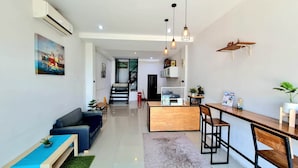Een moderne woonkamer met een bank, salontafel en een eethoek met een houten tafel en stoelen. Er is een trap die naar een hoger niveau leidt, en een schilderij aan de muur.