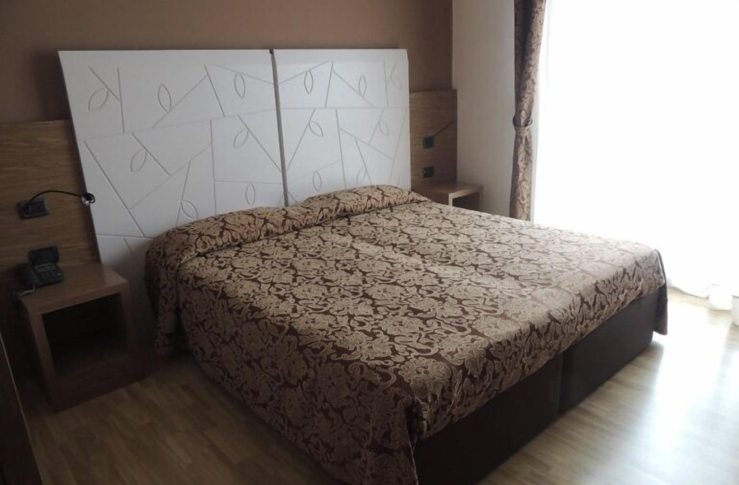 Ein Schlafzimmer mit einem gemusterten Bettbezug, einem hölzernen Kopfende mit geometrischen Mustern, einem Nachttisch mit einem Telefon und einem bei dem Bett stehenden Tisch.
