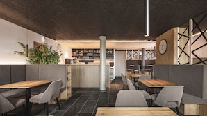 A modern restaurant interior with wooden tables and grey upholstered booths, a bar area with shelves of bottles, and a large clock on the wall. - Hotel Fernblick (Bressanone)