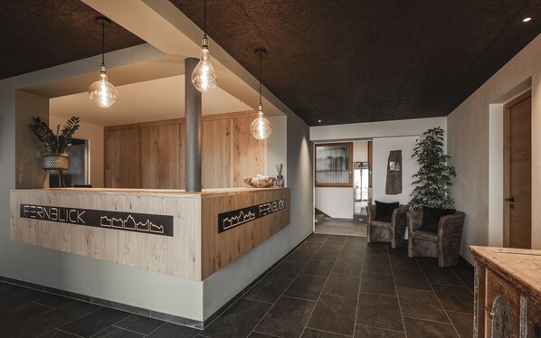A reception area with a wooden counter, pendant lighting, and a view into a living area with a couch and a plant. - Hotel Fernblick (Bressanone)