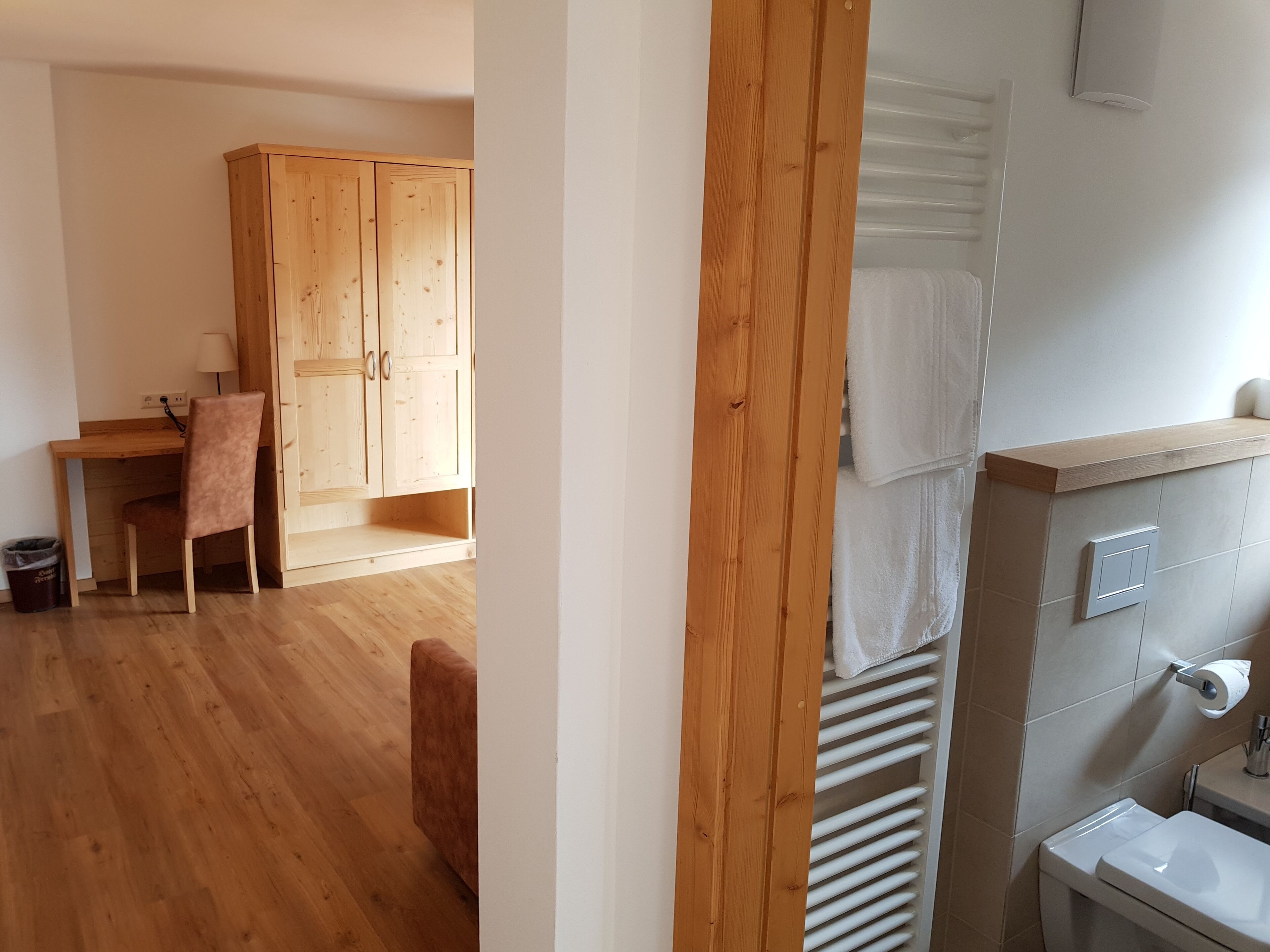 Une pièce comprenant un bureau, une chaise et une armoire en bois. On y trouve également une salle de bain avec des toilettes, un lavabo et un porte-serviettes.