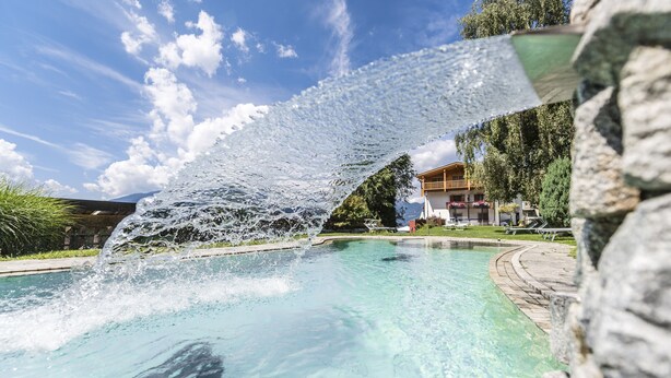 A clear pool with a water feature, surrounded by a stone deck and greenery, with a building in the background. - Hotel Fernblick (Bressanone)