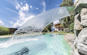 A clear pool with a water feature, surrounded by a stone deck and greenery, with a building in the background. - Hotel Fernblick (Bressanone)