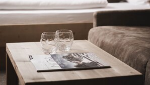 A hotel room with a wooden bed, a wooden nightstand with two glasses and a magazine, and a window with curtains. - Hotel Fernblick (Bressanone)