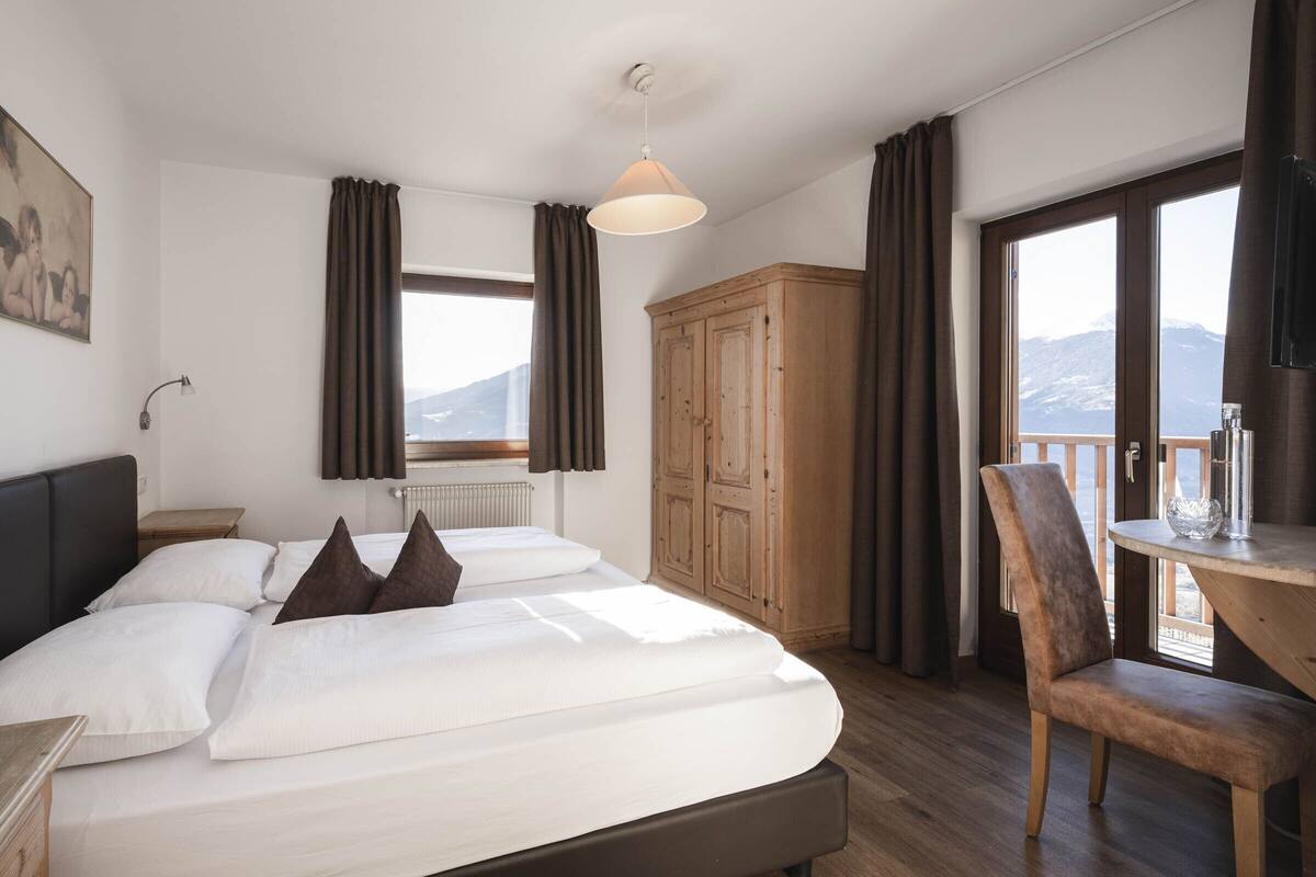 Ein Schlafzimmer mit Bett, einem Holzschrank, einem Schreibtisch mit Stuhl, einem Fenster mit Vorhängen und Blick auf Berge.
