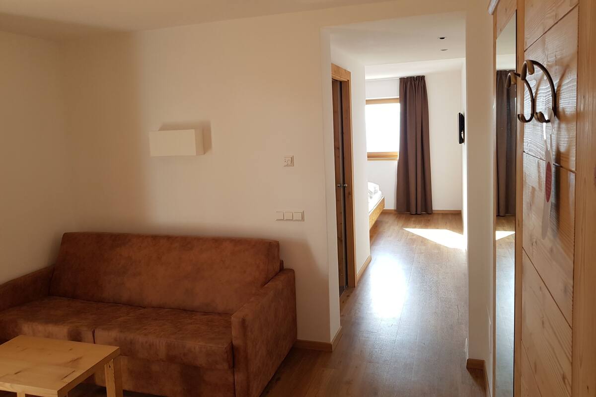 Ein Wohnzimmer mit einem braunen Sofa, einem Holztisch und einem Flur, der zu einem Schlafzimmer mit einem Bett und Vorhängen führt.