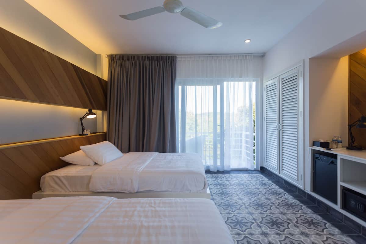 Une chambre d’hôtel avec deux lits, un ventilateur de plafond, un bureau avec une lampe et une vue sur l’extérieur grâce à de grandes fenêtres.