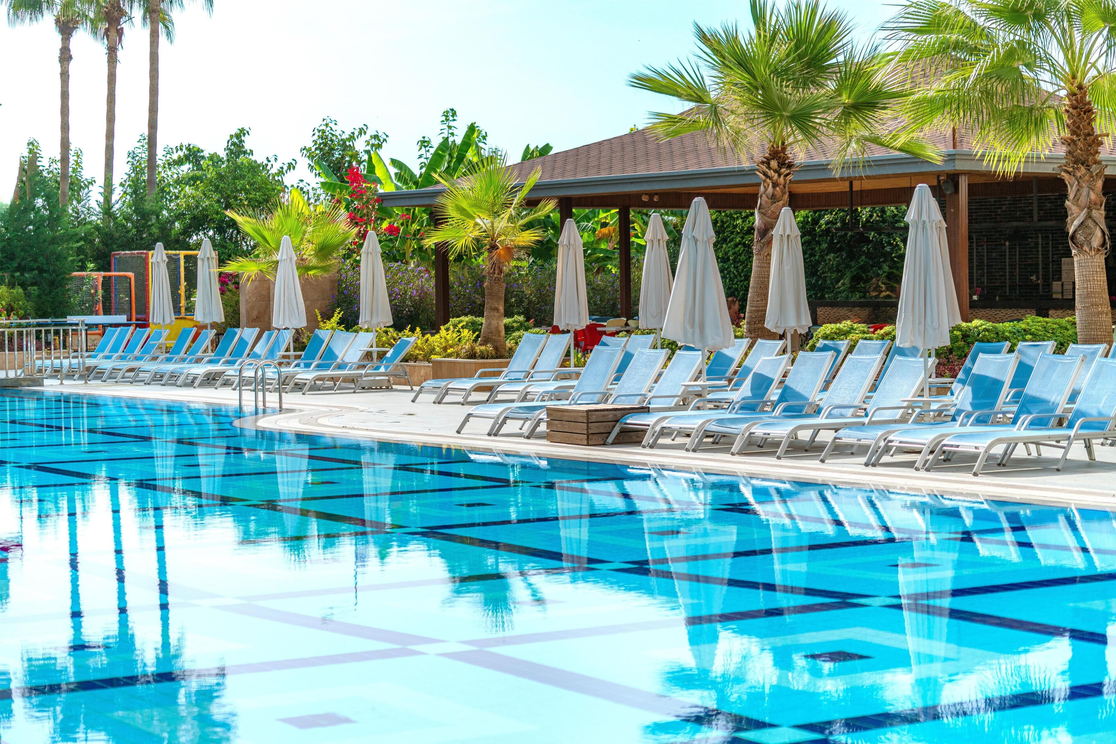Una piscina con carriles azules, rodeada de sillas de descanso y sombrillas, con palmeras y un edificio al fondo.