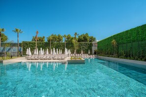 Piscina azul clara con sillas de descanso y sombrillas, rodeada de follaje y un muro alto.