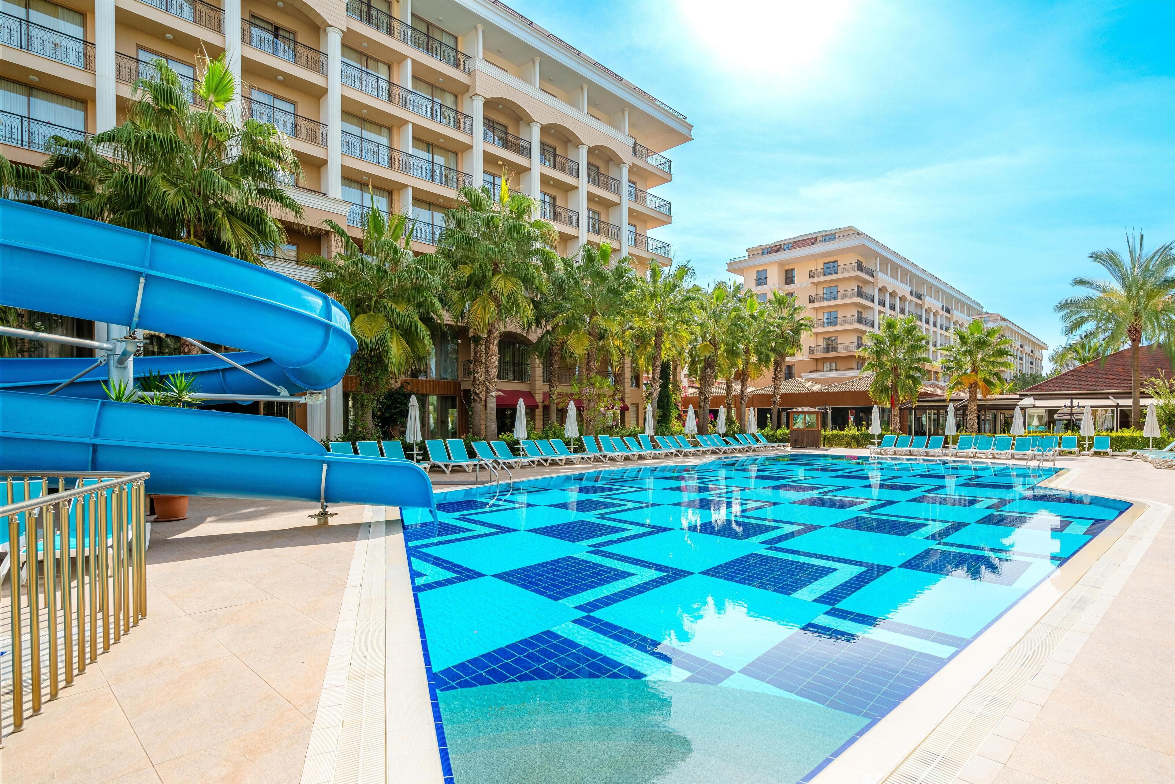 A hotel pool with a blue water slide, palm trees, and lounge chairs.