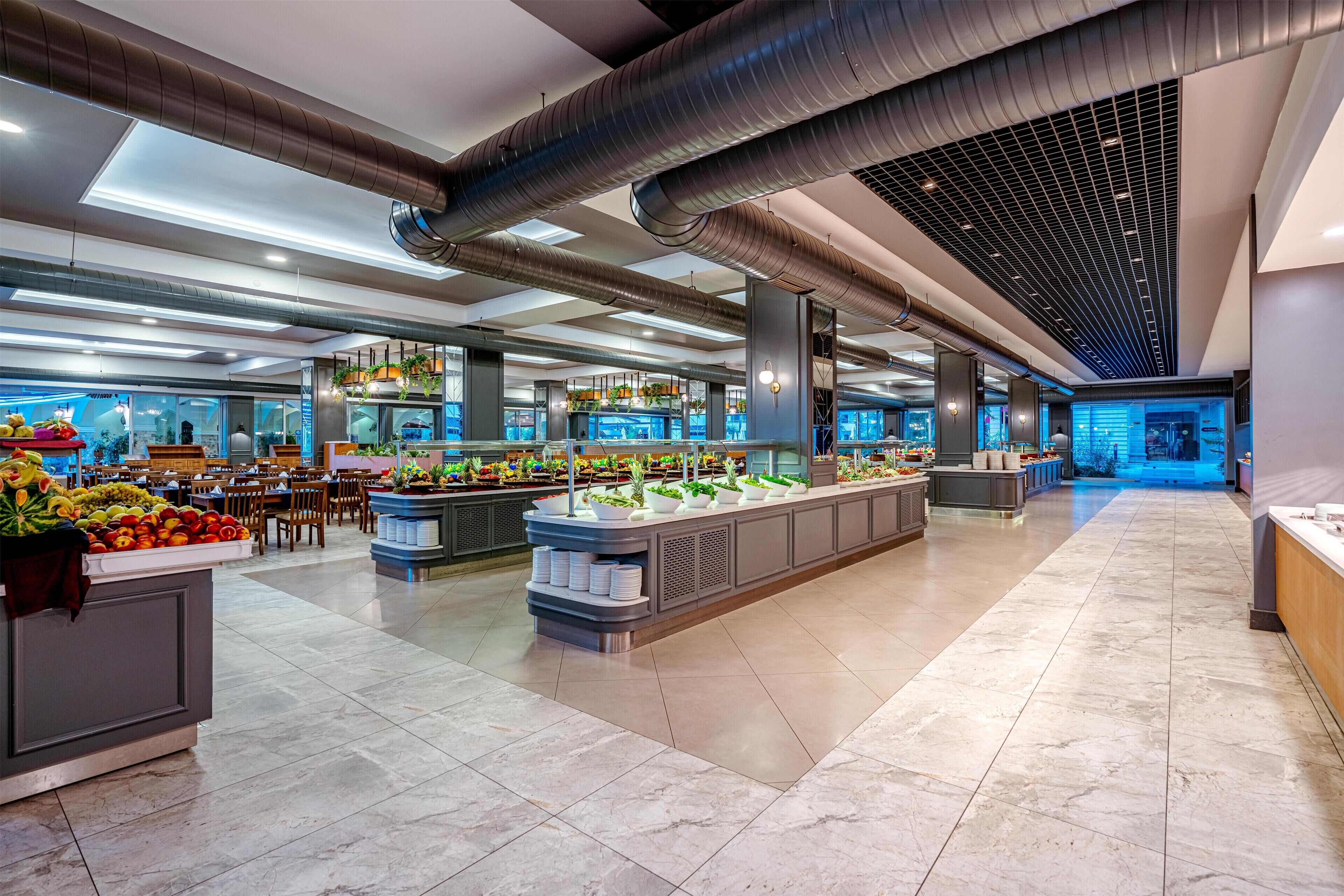 A spacious hotel lobby with a central island featuring a fruit display, multiple seating areas, and a modern ceiling design with exposed ductwork.