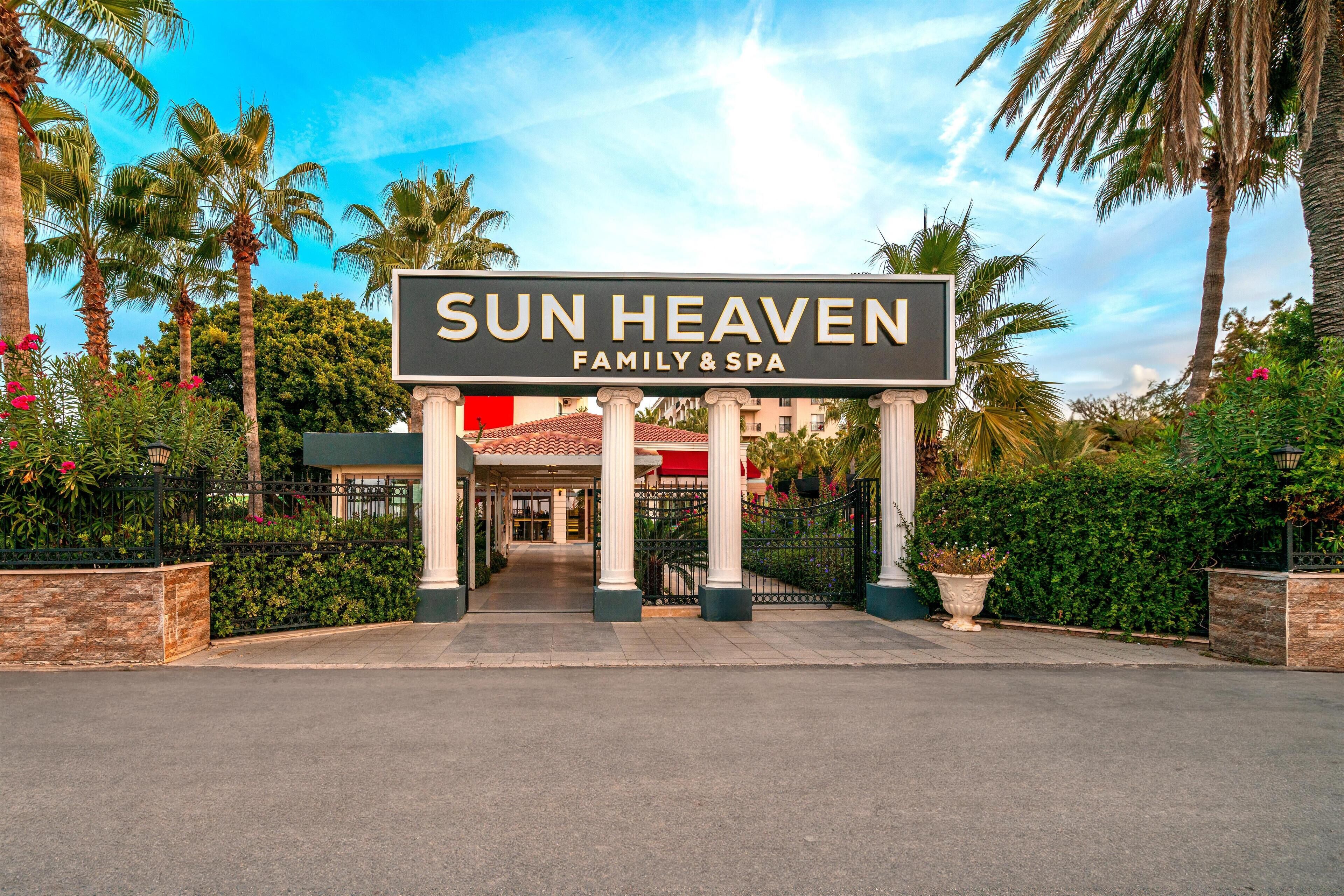 La entrada a Sun Heaven Family & Spa, con un letrero llamativo, palmeras y un camino bien cuidado que lleva al edificio.