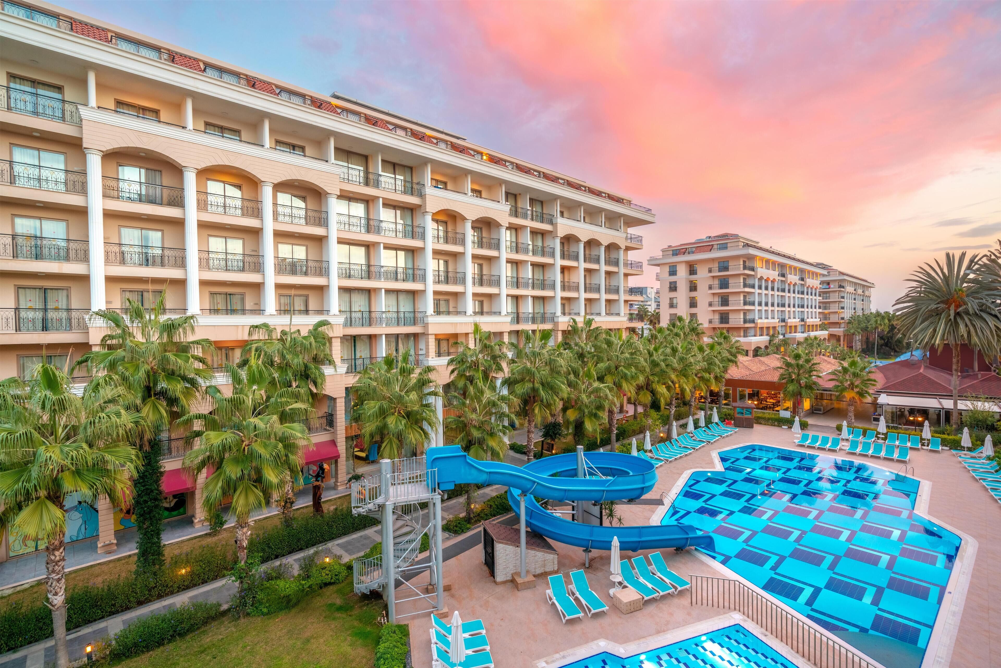A hotel with multiple balconies, a pool area with lounge chairs, and palm trees.