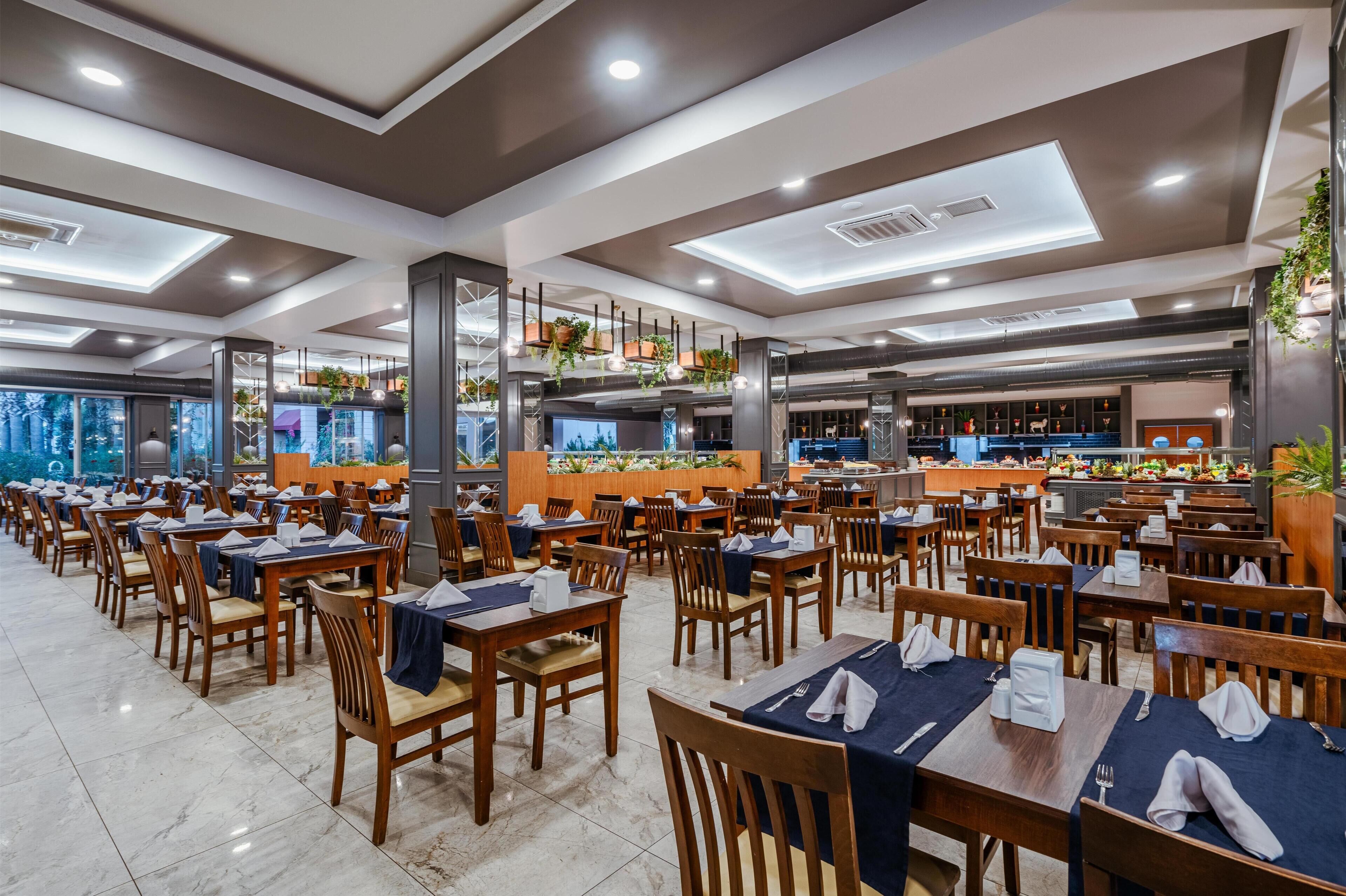A spacious restaurant with multiple dining tables and chairs, a central counter with a display of food items, and a ceiling with recessed lighting.
