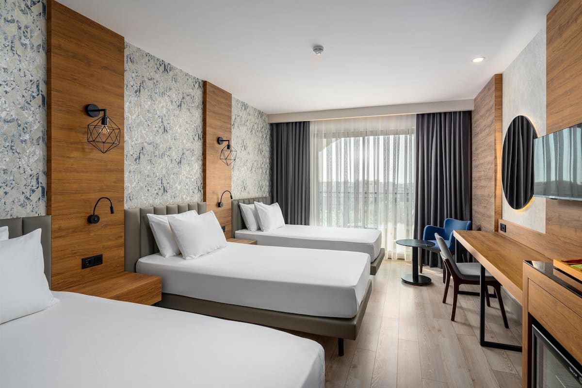 A hotel room with two beds, a desk, and a chair. There is a large window with curtains, a flat-screen TV mounted on the wall, and a wooden accent wall.