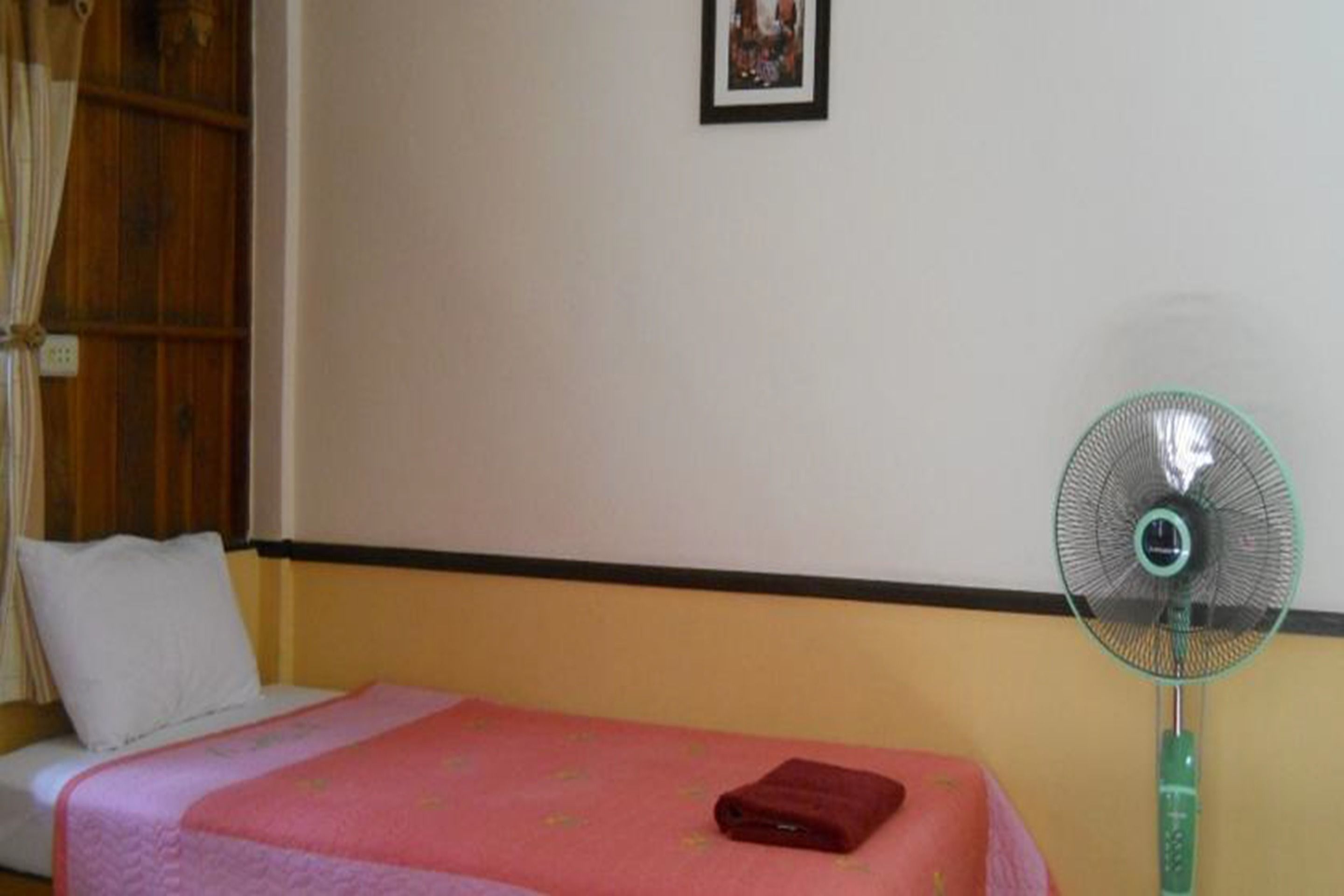 Une chambre avec un lit, une couvre-lit rose, un oreiller blanc, un ventilateur vert sur pied et un petit tableau encadré accroché au mur.