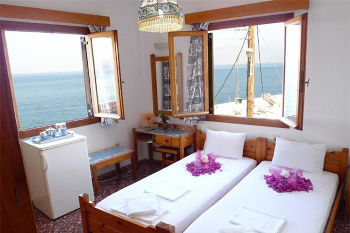 Ein Zimmer mit zwei Betten, einem Schreibtisch, einer kleinen Kühlbox und großen Fenstern mit Meerblick.