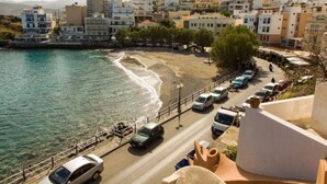A coastal road with parked cars, a beach, and buildings in the background. - Pension Mylos (Agios Nikolaos)