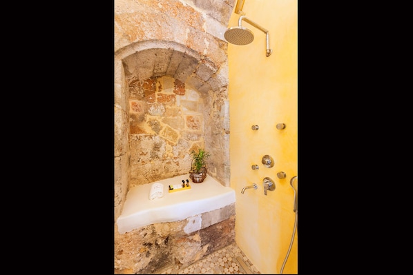 Une salle de bain aux murs en pierre, dotée d’un lavabo intégré, d’une douche et d’une petite étagère où sont rangés les articles de toilette.