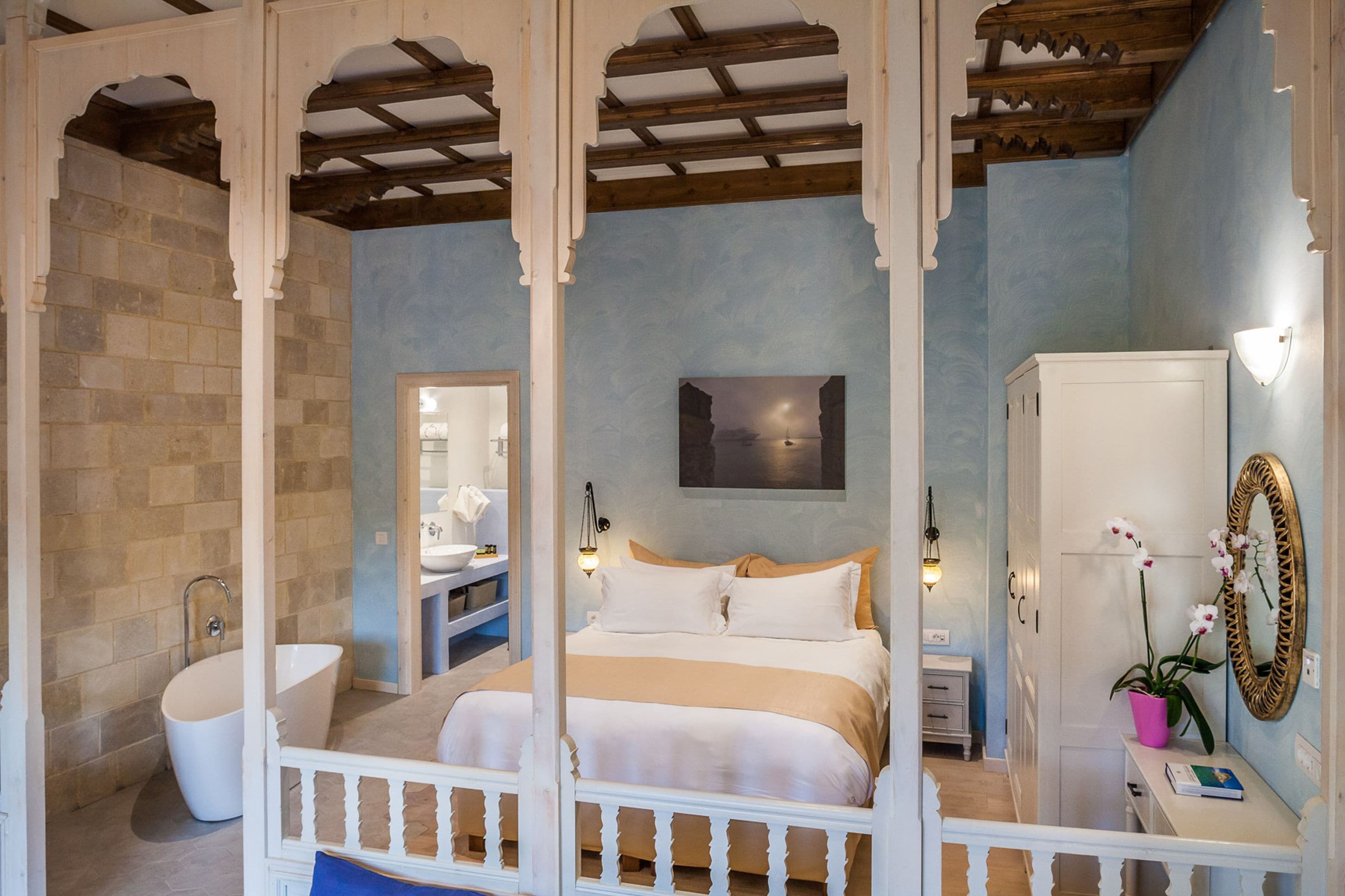 Une chambre à coucher avec un lit, une baignoire blanche, une coiffeuse avec un miroir et un pot d’orchidée.