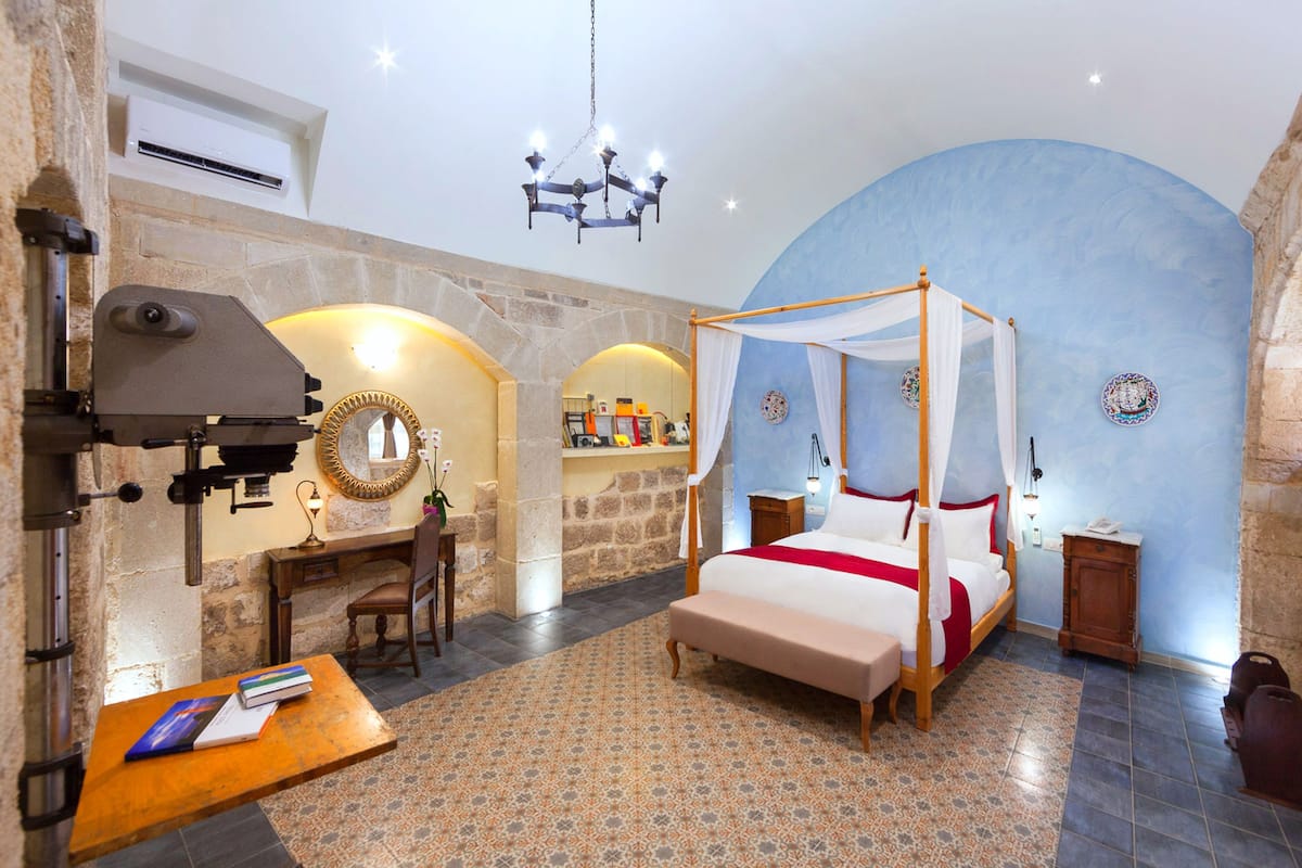A bedroom with a four-poster bed, a desk with a chair, a vanity with a mirror, and a chandelier.