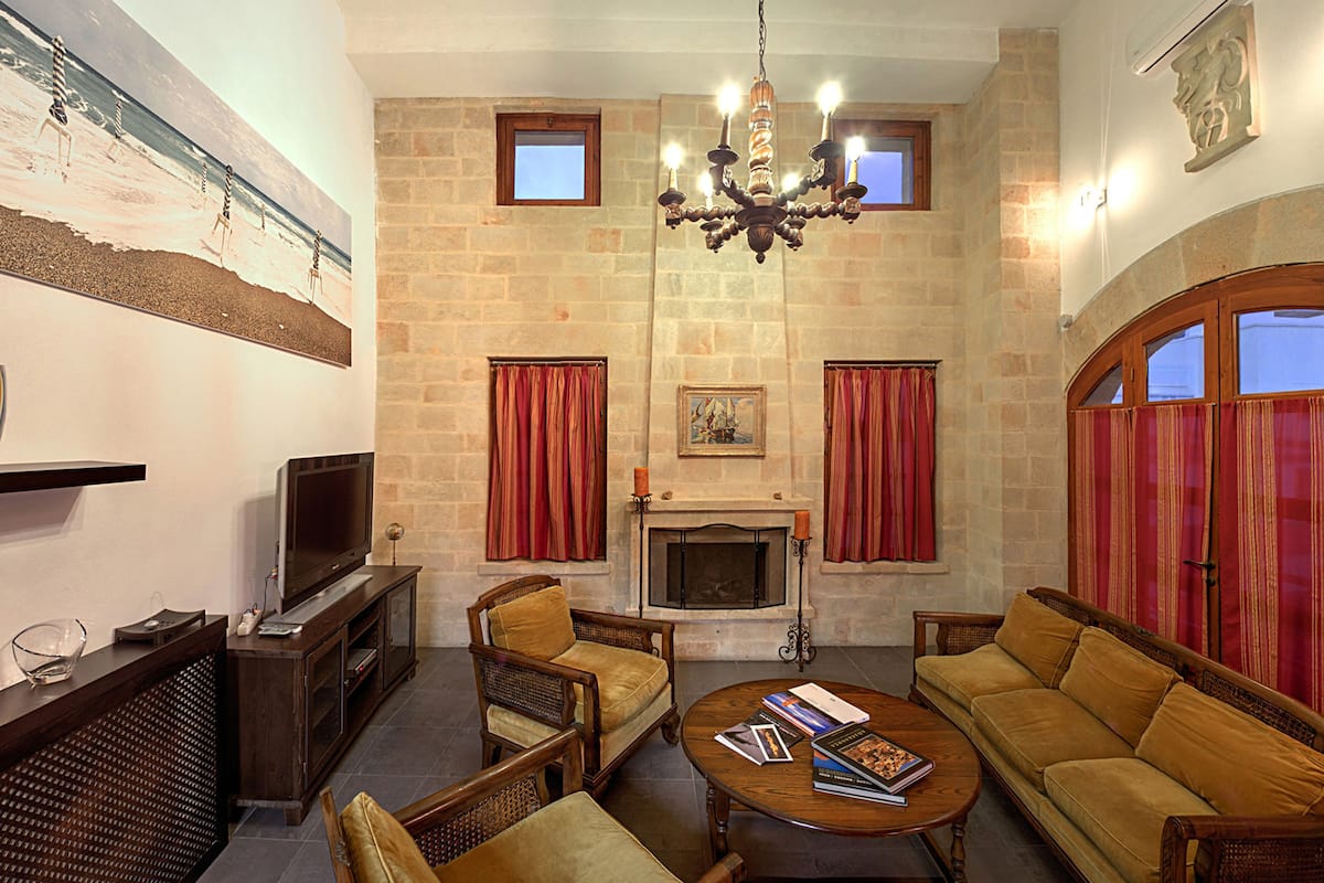 A living room with a fireplace, stone walls, red curtains, and a chandelier.