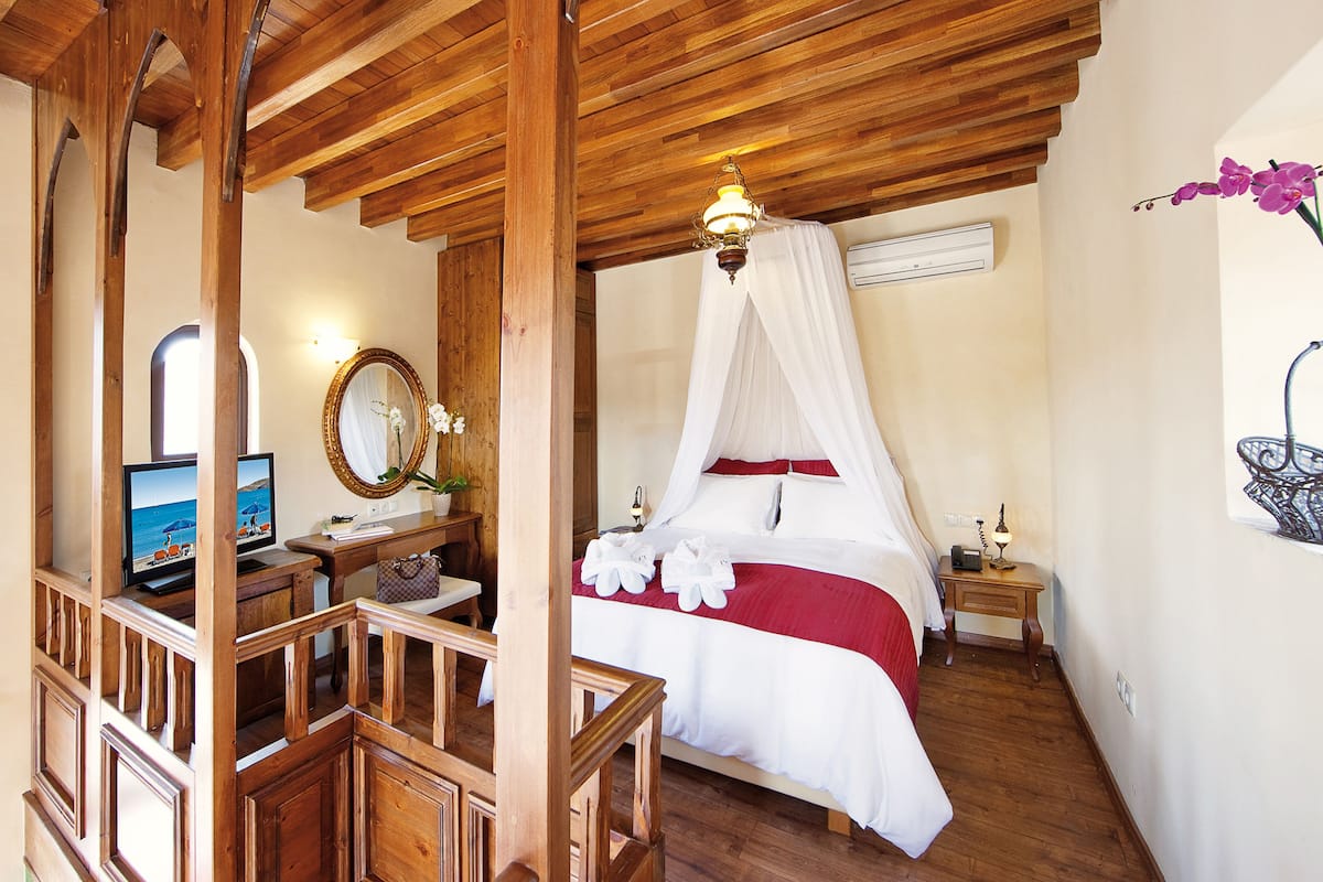 A bedroom with a wooden ceiling, a bed with a canopy, a bedside table with a lamp, a desk with a computer, and a mirror.