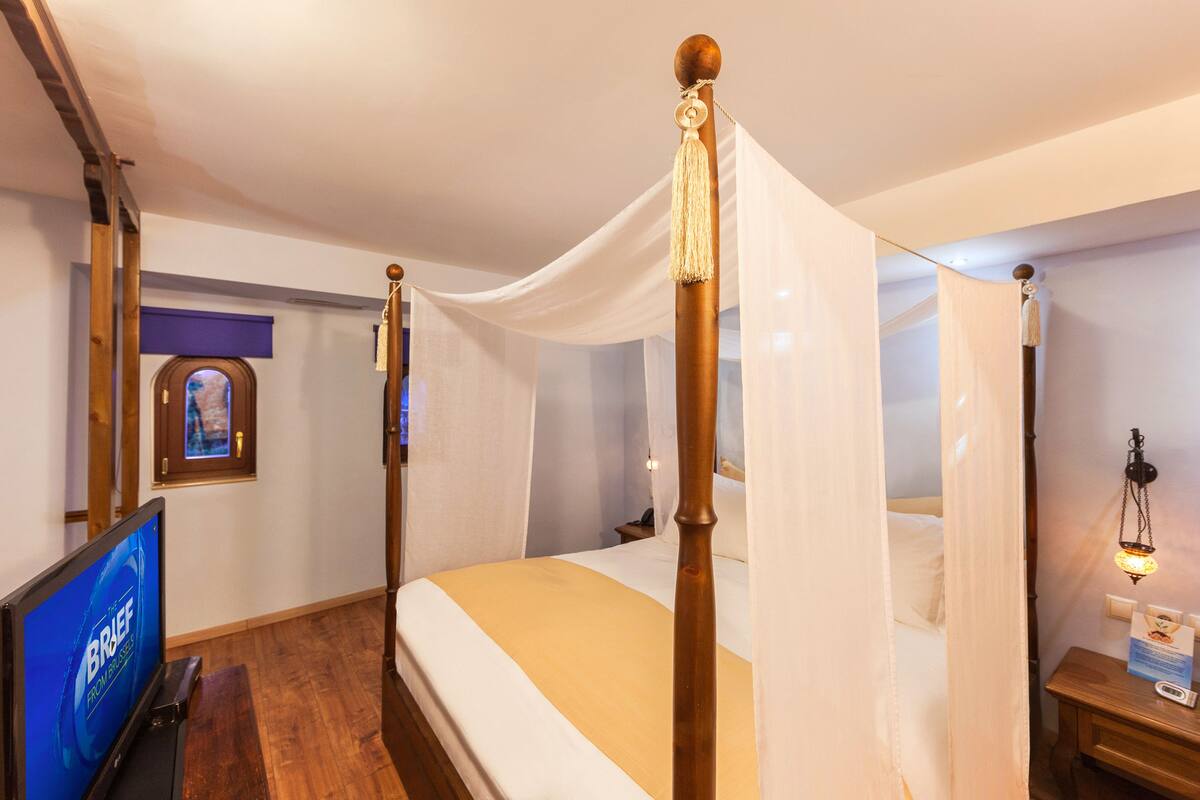 A room with a four-poster bed, a television, a wooden nightstand, and a wall-mounted lamp.