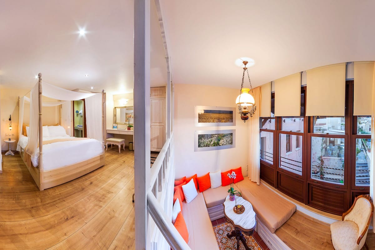 A room with a four-poster bed, a sofa, a small table, and a chair. There is a window with a view of buildings outside.