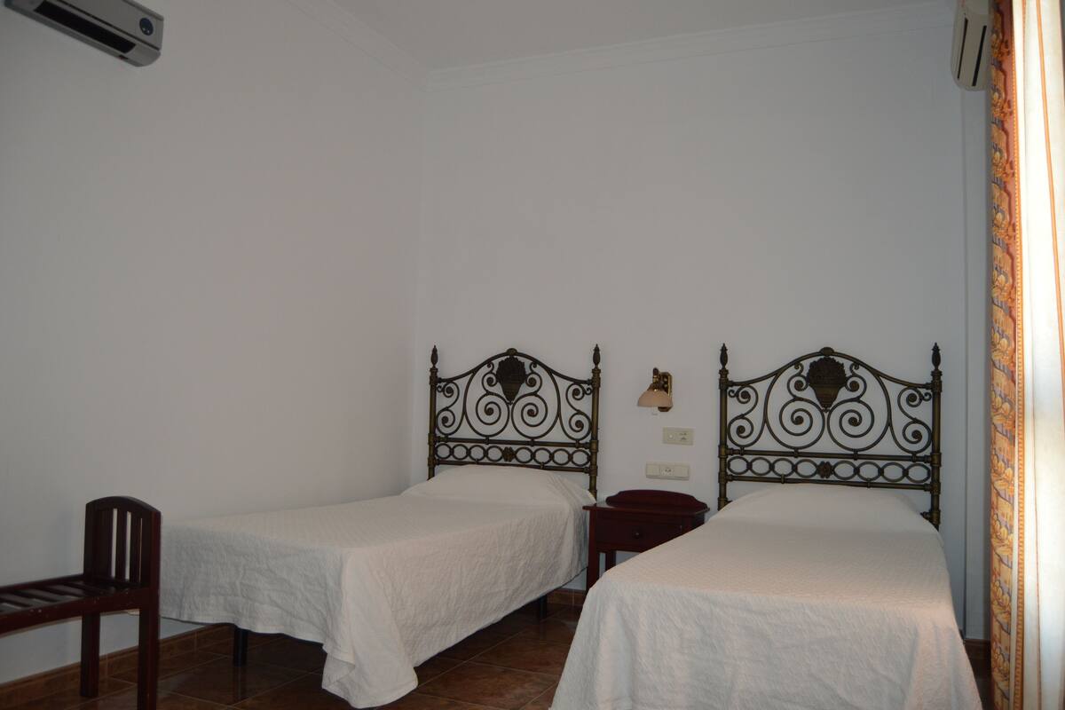 A room with two single beds, a wooden chair, a small table, and a wall-mounted lamp.