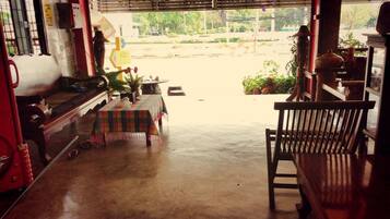 A room with a concrete floor, a wooden bench, a table with a plaid tablecloth, and a view of a street and greenery outside.