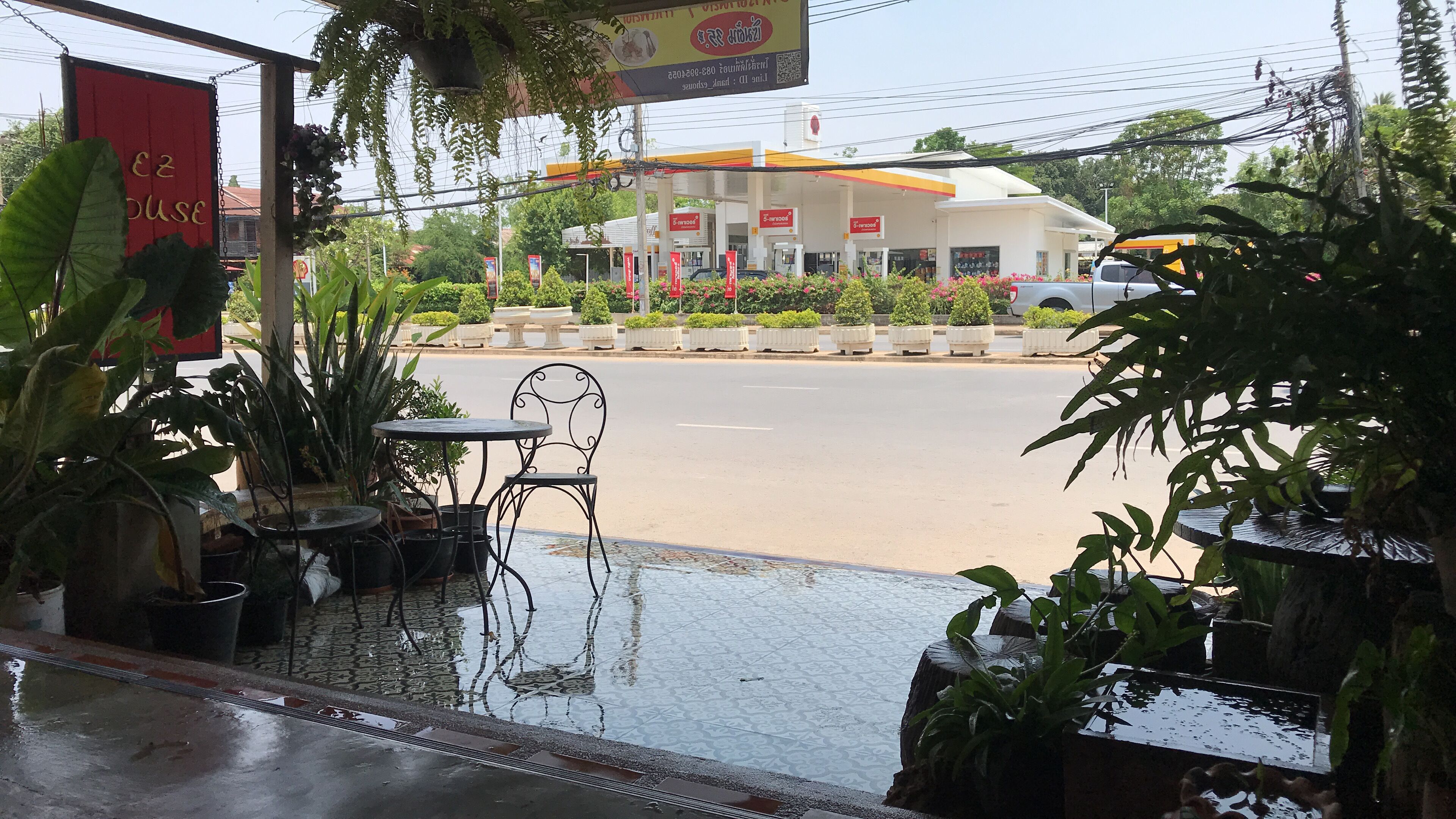 Ein Café am Straßenrand mit Außenbestuhlung, im Hintergrund eine Tankstelle und rundherum Kübelpflanzen.