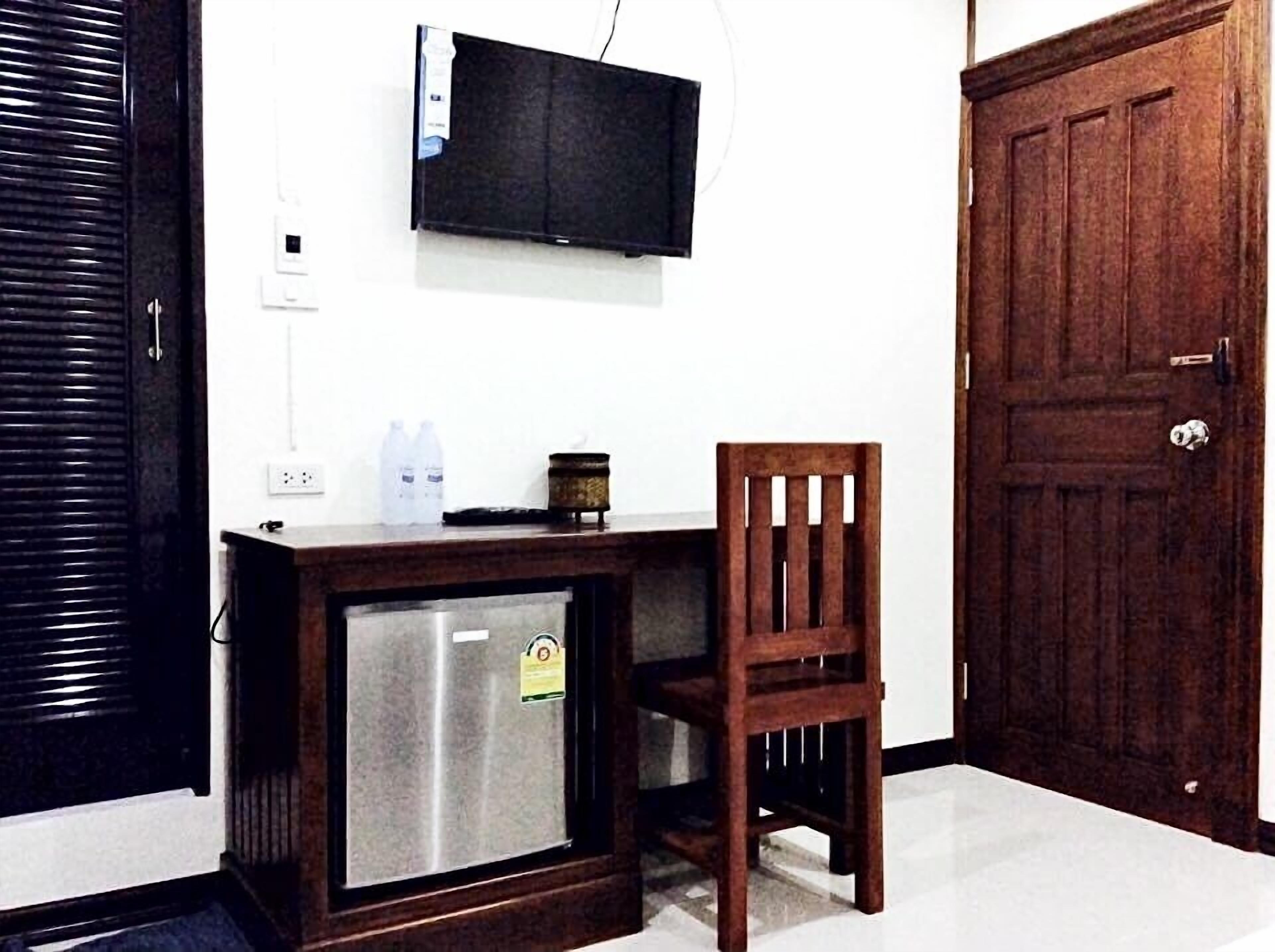 A compact room with a wooden desk, a small refrigerator, a television mounted on the wall, and a wooden door.
