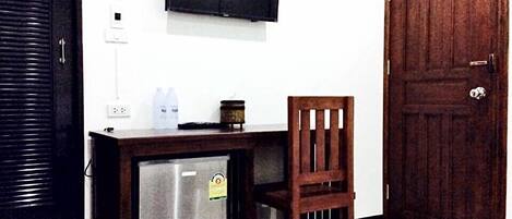 Habitación compacta con escritorio de madera, un refrigerador pequeño, televisor montado en la pared y puerta de madera.