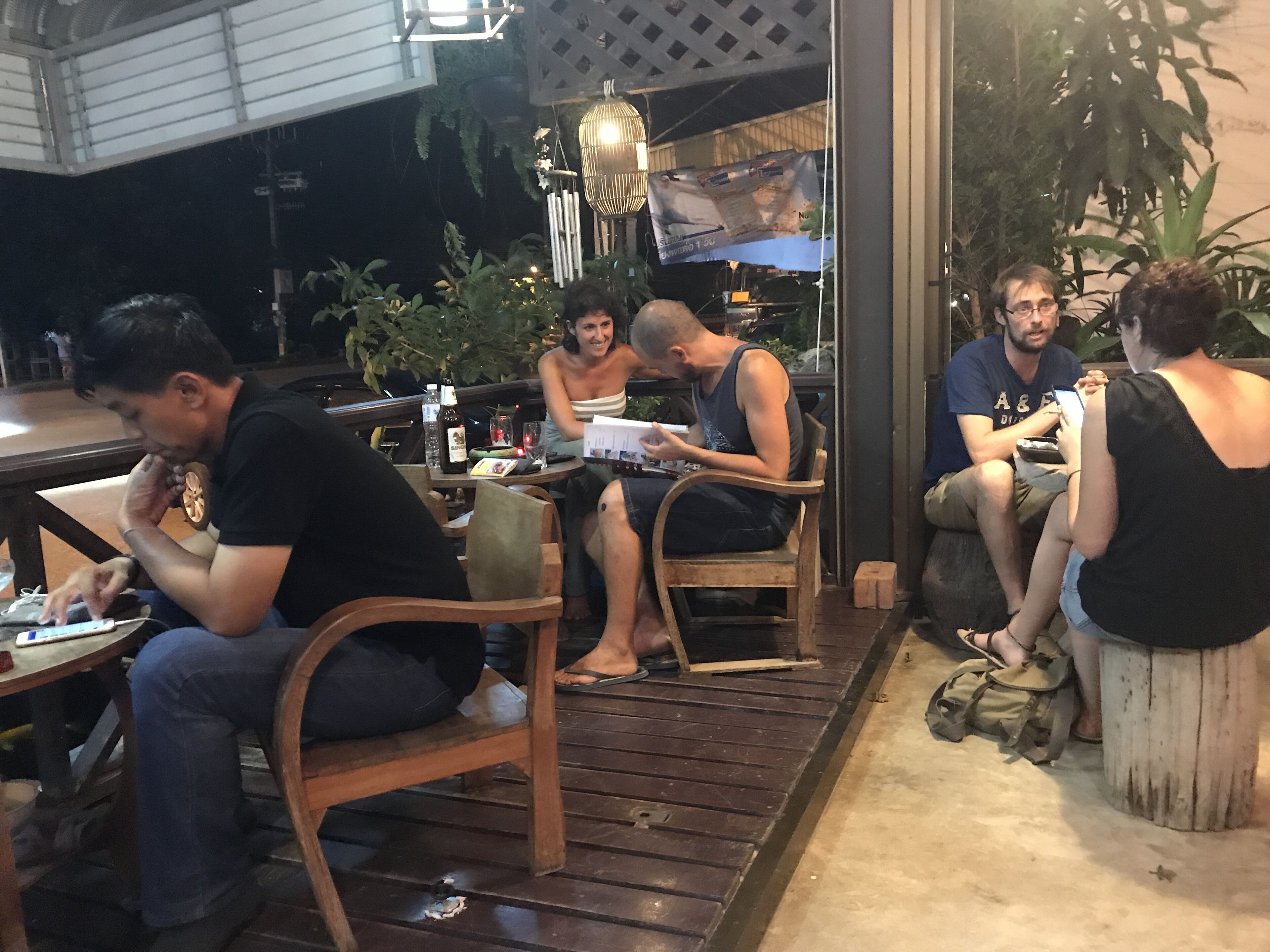 A group of people seated at an outdoor table with a bottle and glasses, surrounded by plants and a decorative light fixture.