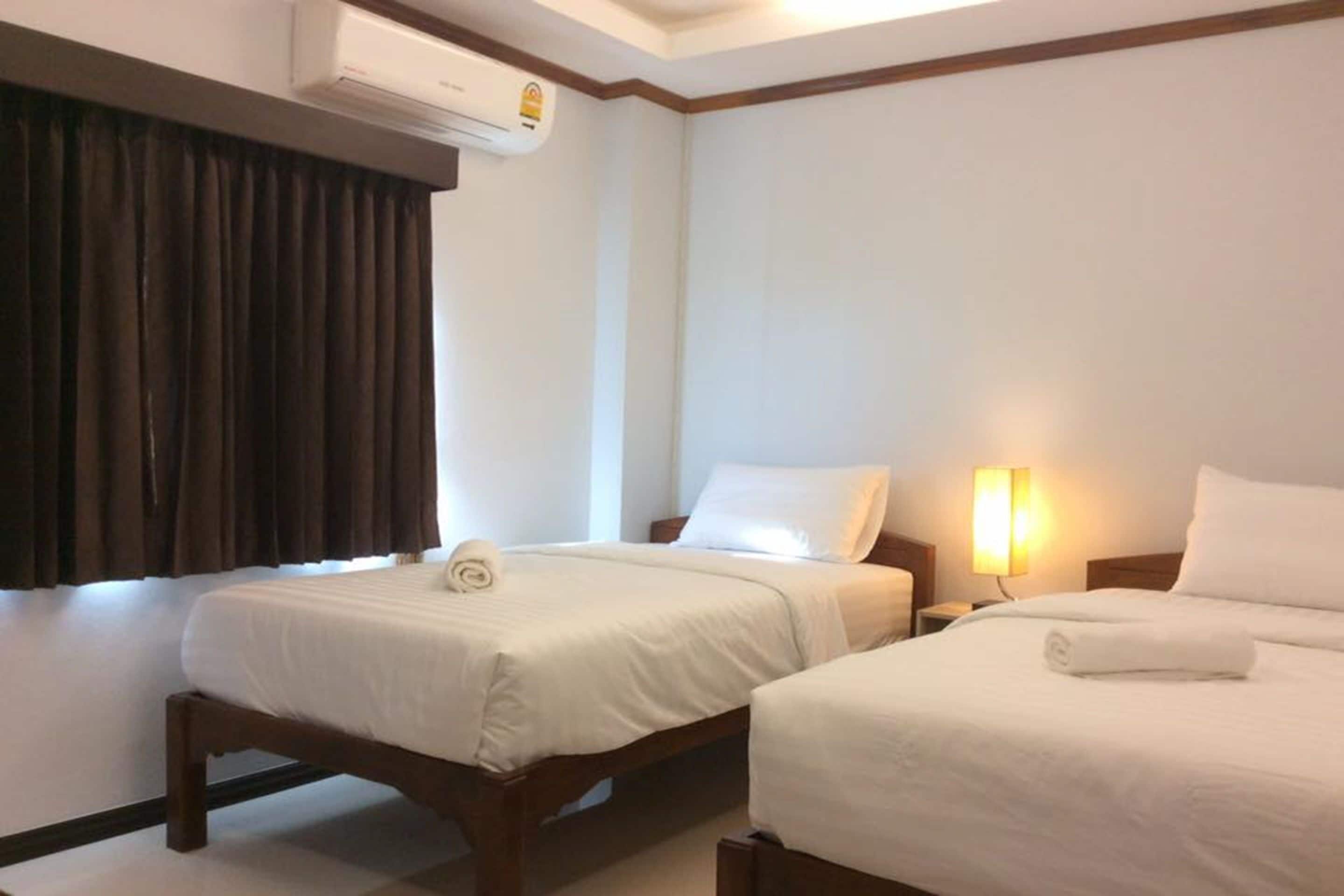 A hotel room with two single beds, a window with curtains, an air conditioning unit, and a bedside lamp.
