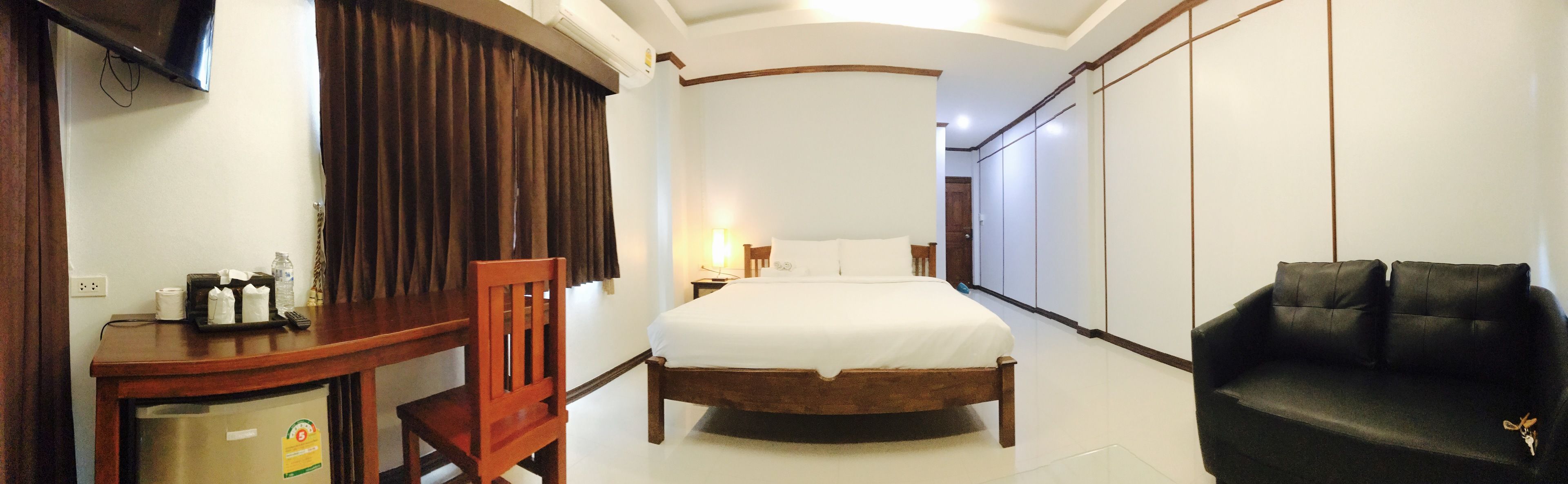 A bedroom with a bed, a wooden chair, a black sofa, a desk with a lamp, and a wardrobe.