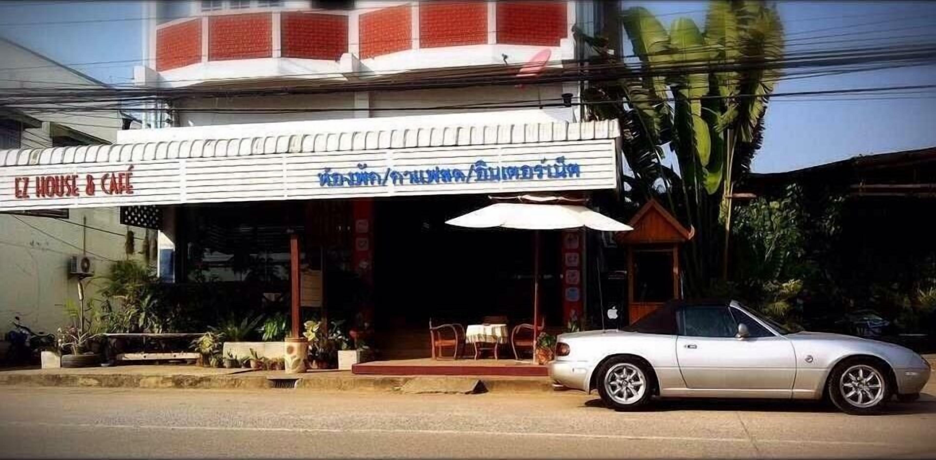 A building with a sign reading "EZ HOUSE & CAFE" and another sign in a different language. There is a white car parked in front.