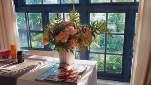 A room with a large window, a white table with a magazine, a vase with flowers, and a white furry rug. - Fawlty Towers Guest House (Livingstone)