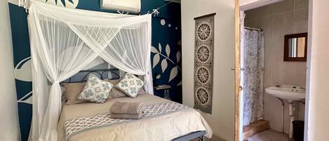 A bedroom with a bed, a mosquito net, a wall-mounted air conditioner, and a bathroom with a sink and mirror.