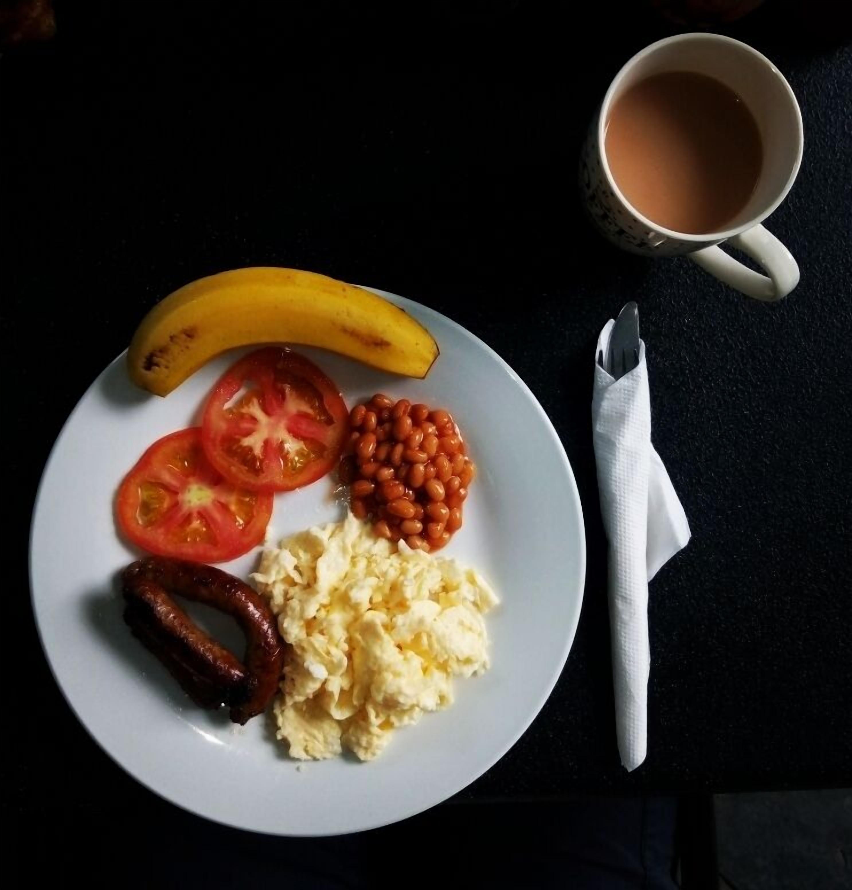 Une assiette contenant une banane, des tranches de tomate, des haricots cuits, des œufs brouillés et des saucisses, accompagnée d’une tasse de thé et d’une serviette en papier, le tout posé sur une surface sombre.