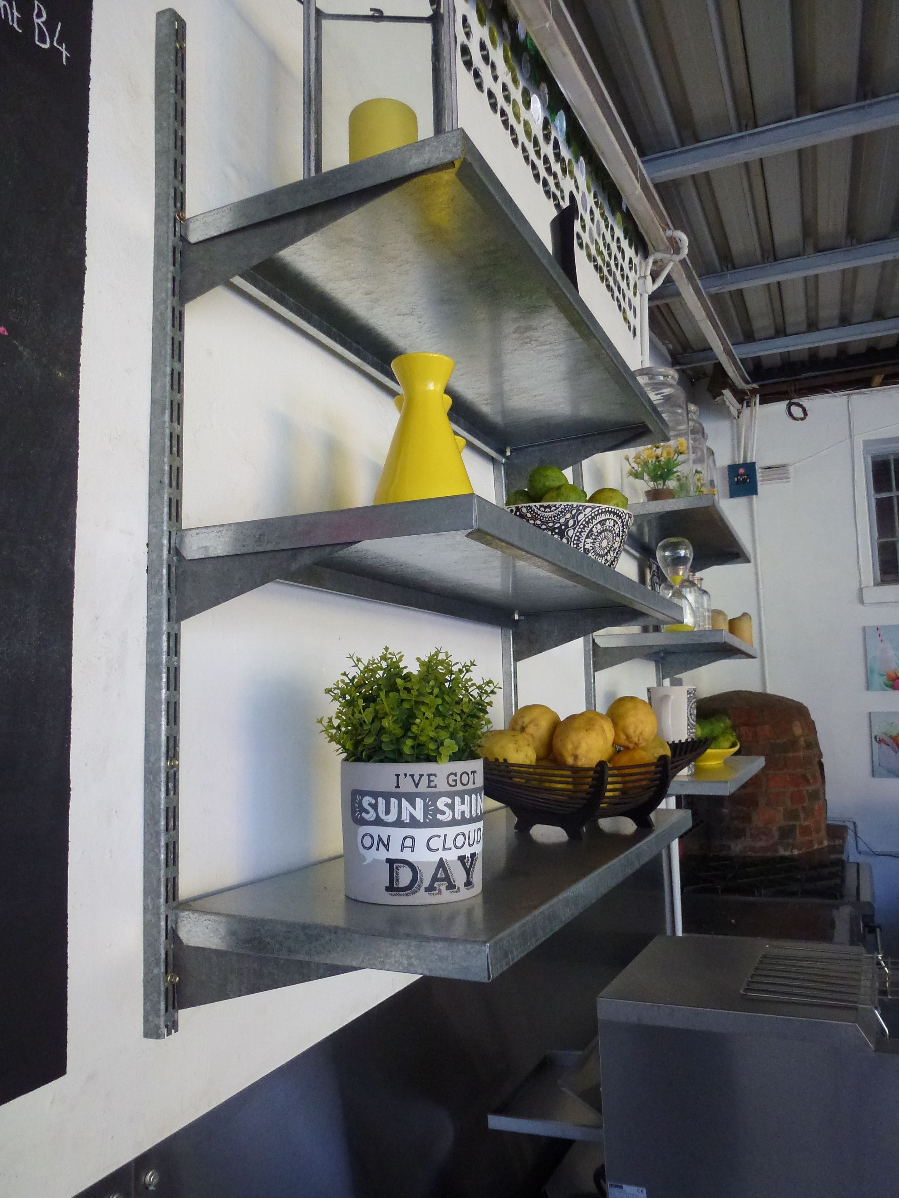 A kitchen with metal shelves holding various items including a yellow vase, a potted plant, and a bowl of fruit.