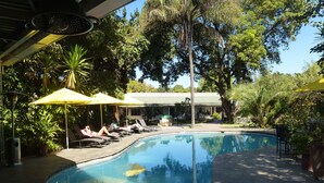 A swimming pool with lounge chairs and umbrellas, surrounded by trees and a building in the background. - Fawlty Towers Guest House (Livingstone)