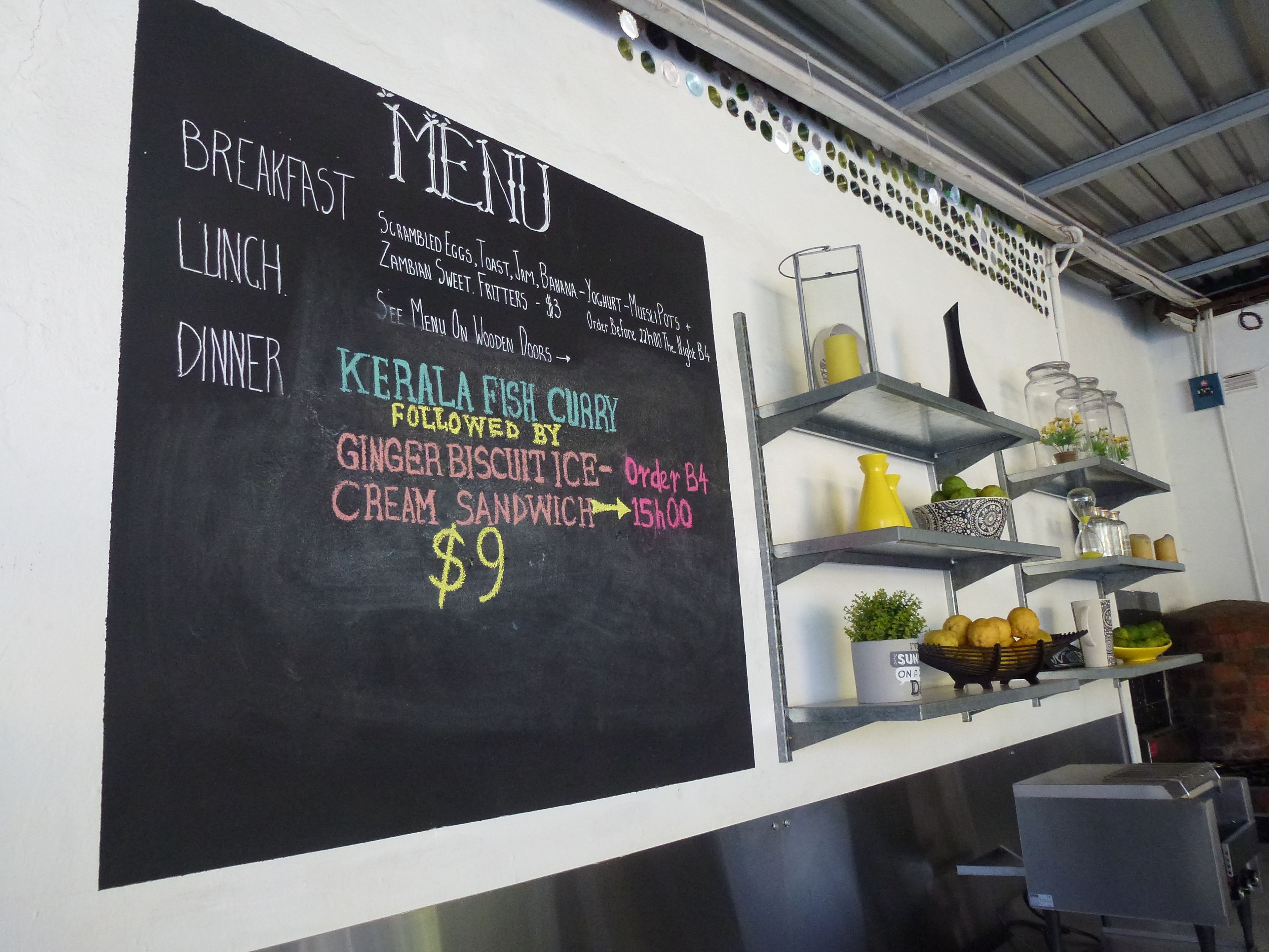 A chalkboard menu with breakfast, lunch, and dinner options, including Kerala fish curry and ginger biscuit ice cream sandwich.