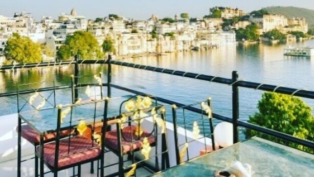 A terrace with a table and chairs overlooking a river and a town. - Hotel Hanuman Ghat (Udaipur)