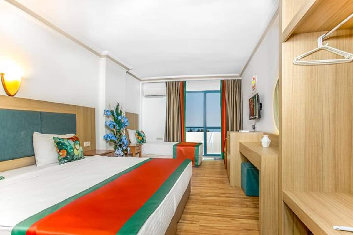 A hotel room with a bed, a desk, a chair, a small table, a plant, and a view of the outside.