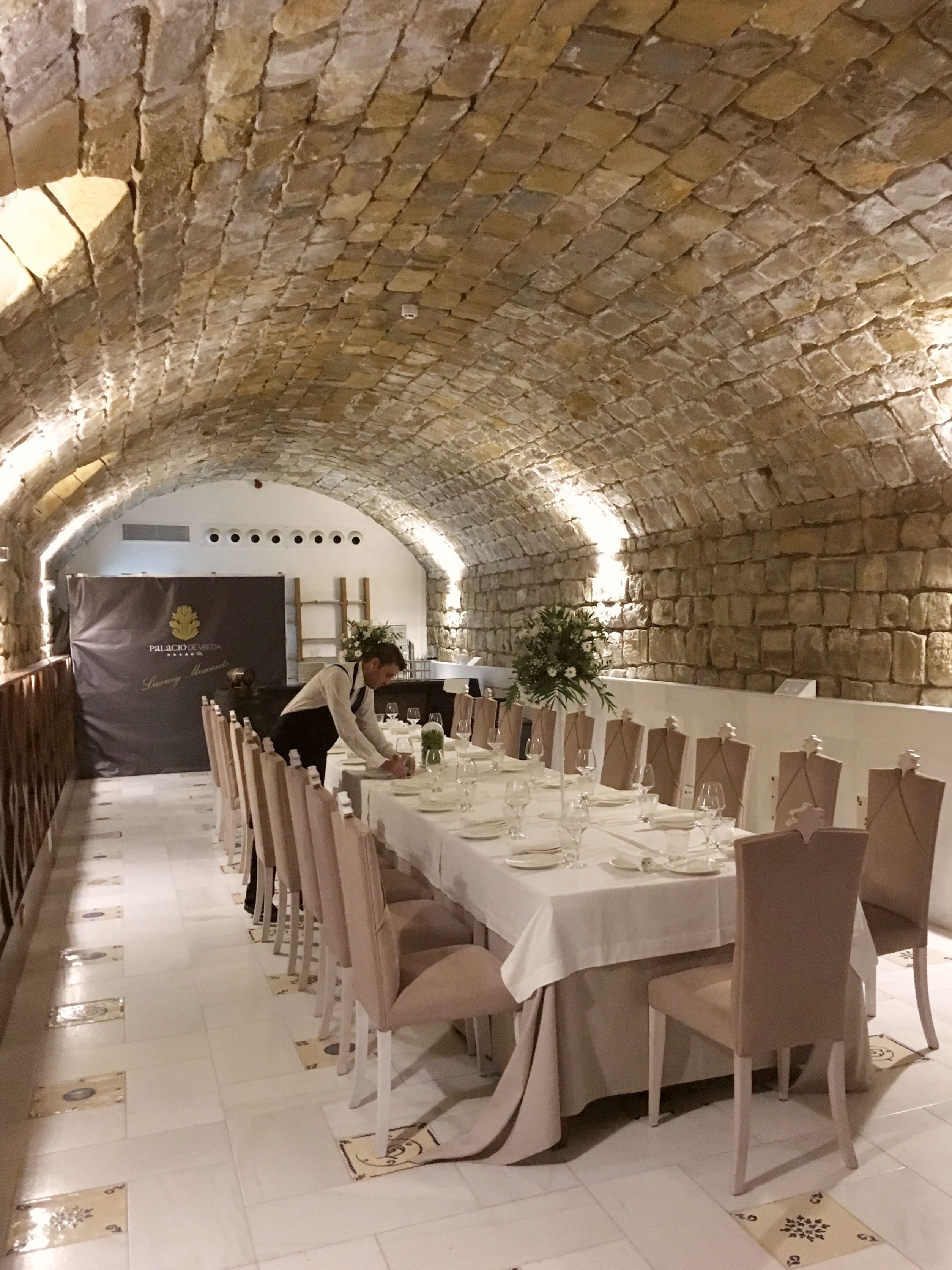 A long dining table set with chairs, a waiter, and a banner in a stone-walled room.