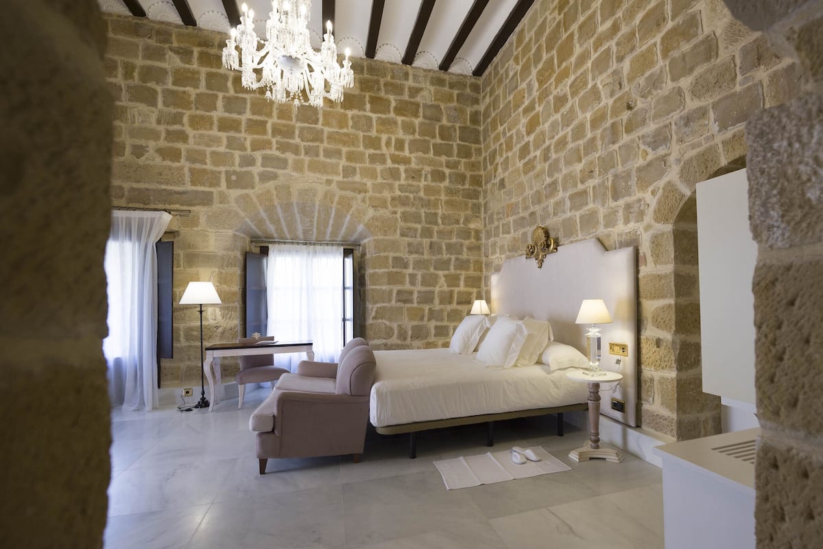 Un dormitorio con pared de piedra, cama, silla, escritorio y candelabro.