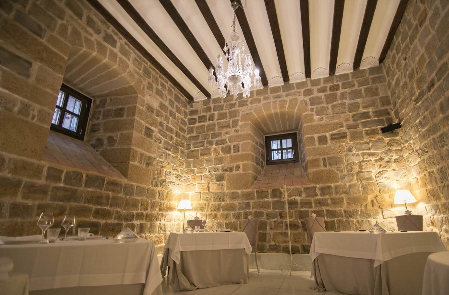 Una habitación de paredes de piedra con dos mesas, cada una con mantel y candelabro.  Un candelabro cuelga del techo y hay una ventana pequeña con vistas.