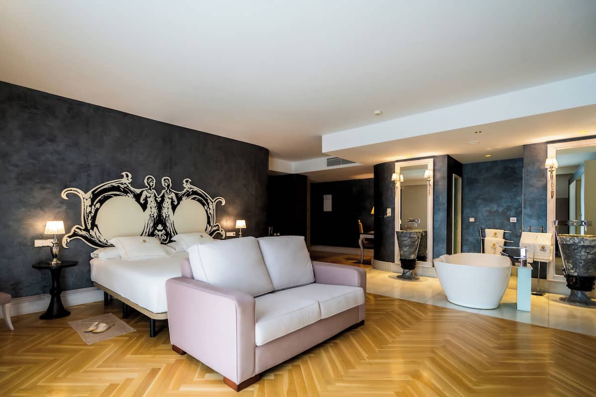 Un dormitorio moderno con una cama grande, un sofá rosa, una bañera blanca y una pared de acento oscura.